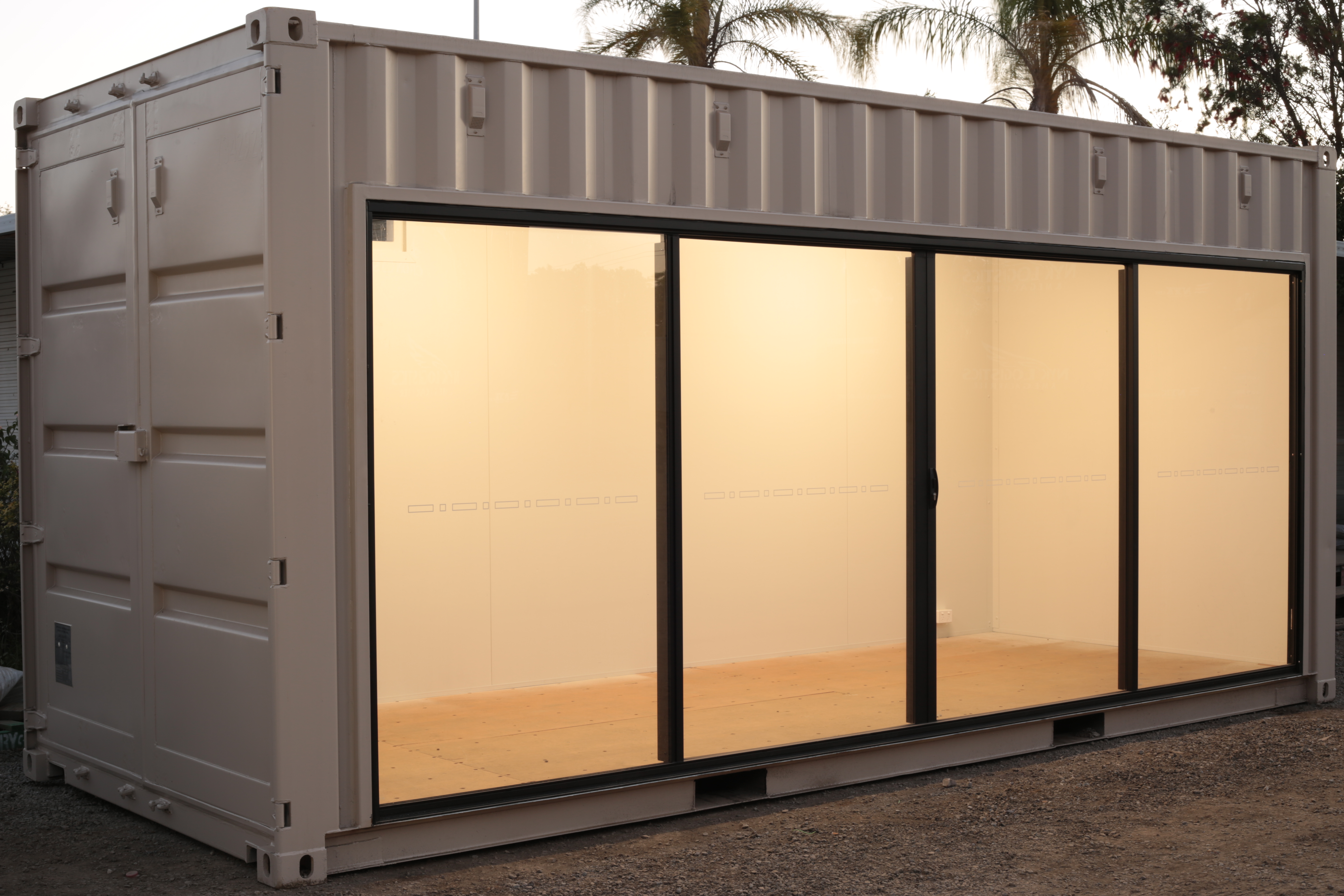 A cream 20ft shipping container with a glass sliding door the lengh of one side. There is warm light emanating from the interior.