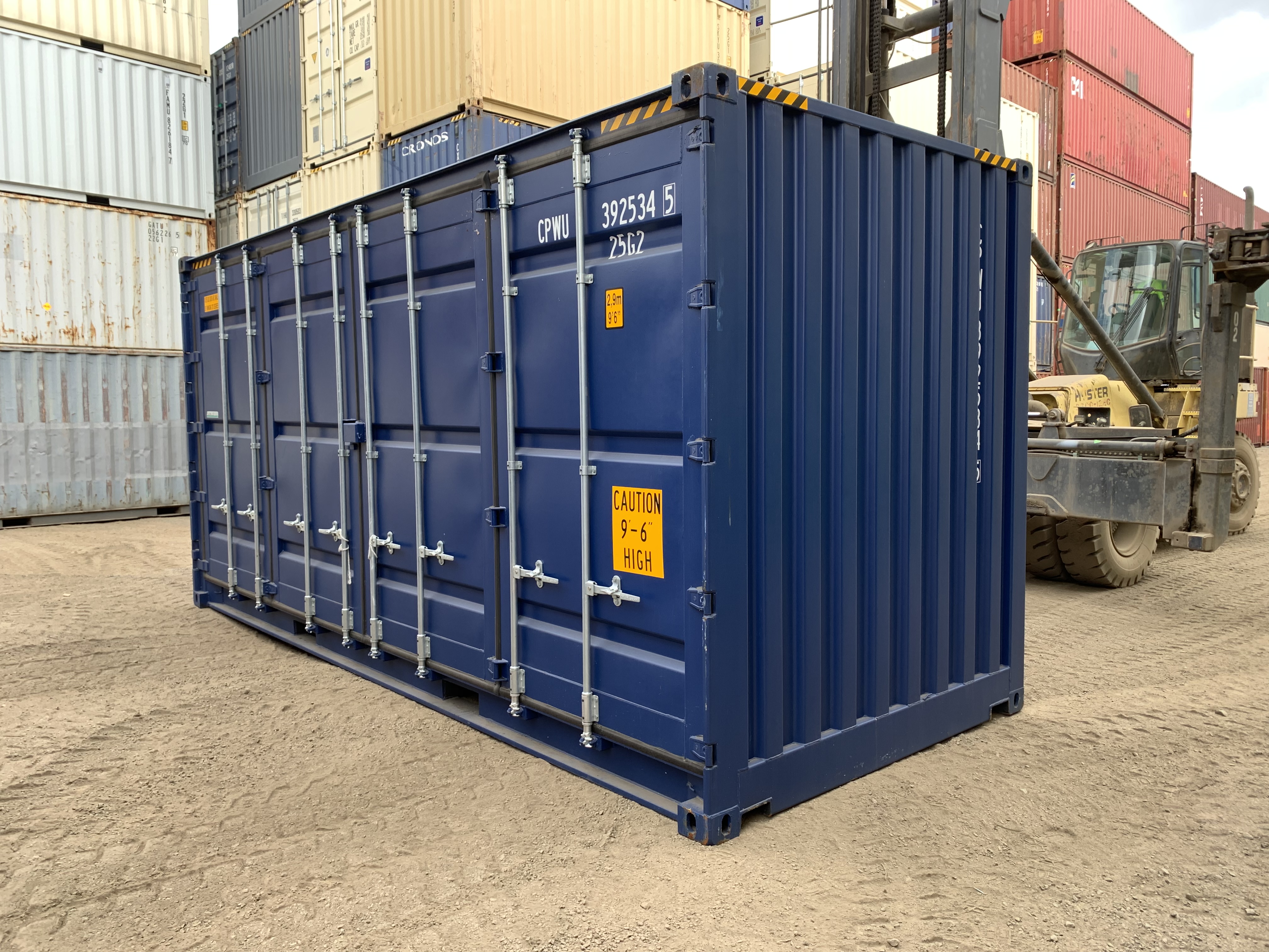 A dark blue side opening twenty-foot shipping container sits in a depot. A container handler and stack of shipping containers are visible in the background.