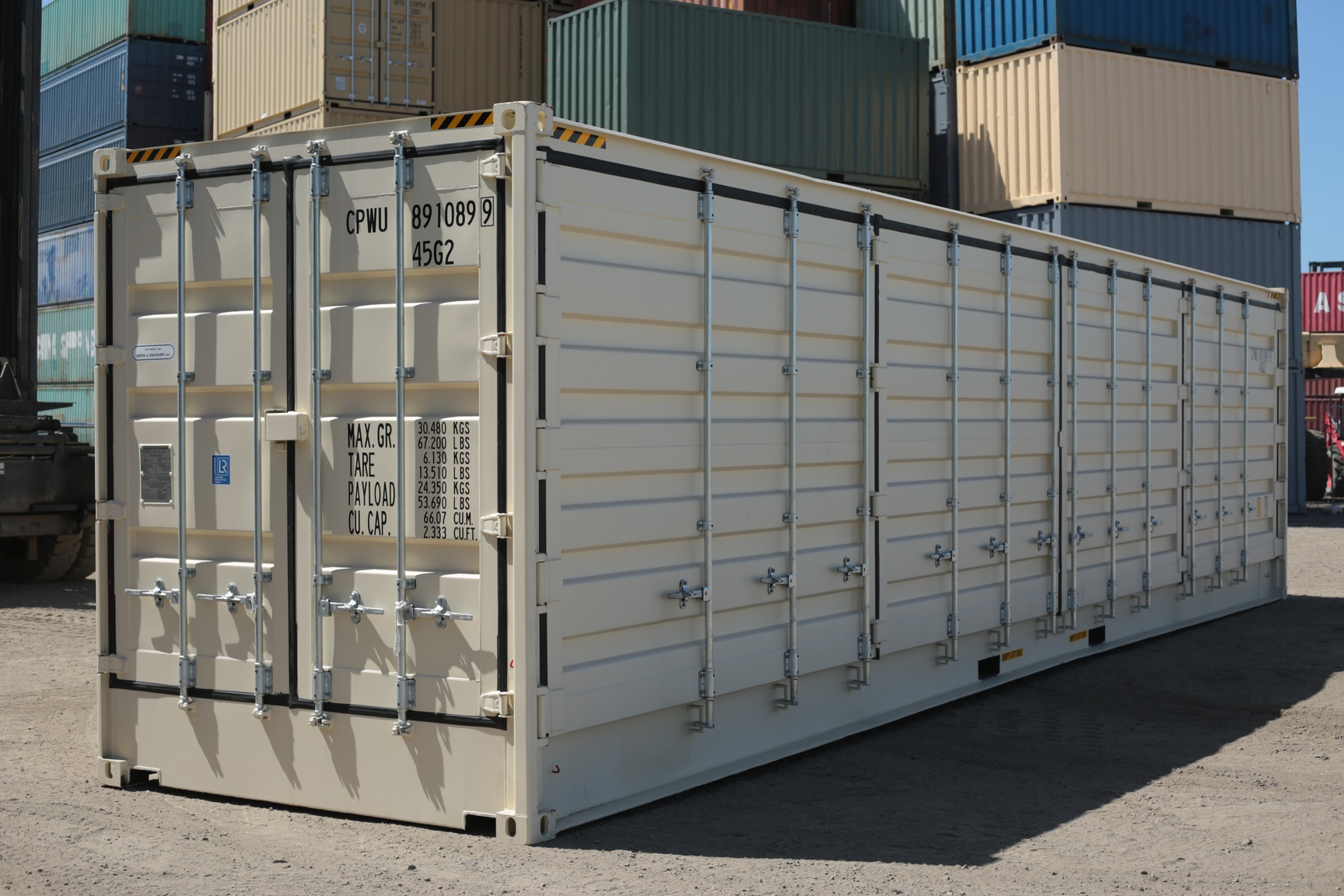 A cream coloured fourty foot shipping container sits in a dusty depot. The container has doors visible down its side in addition to the doors on the front.